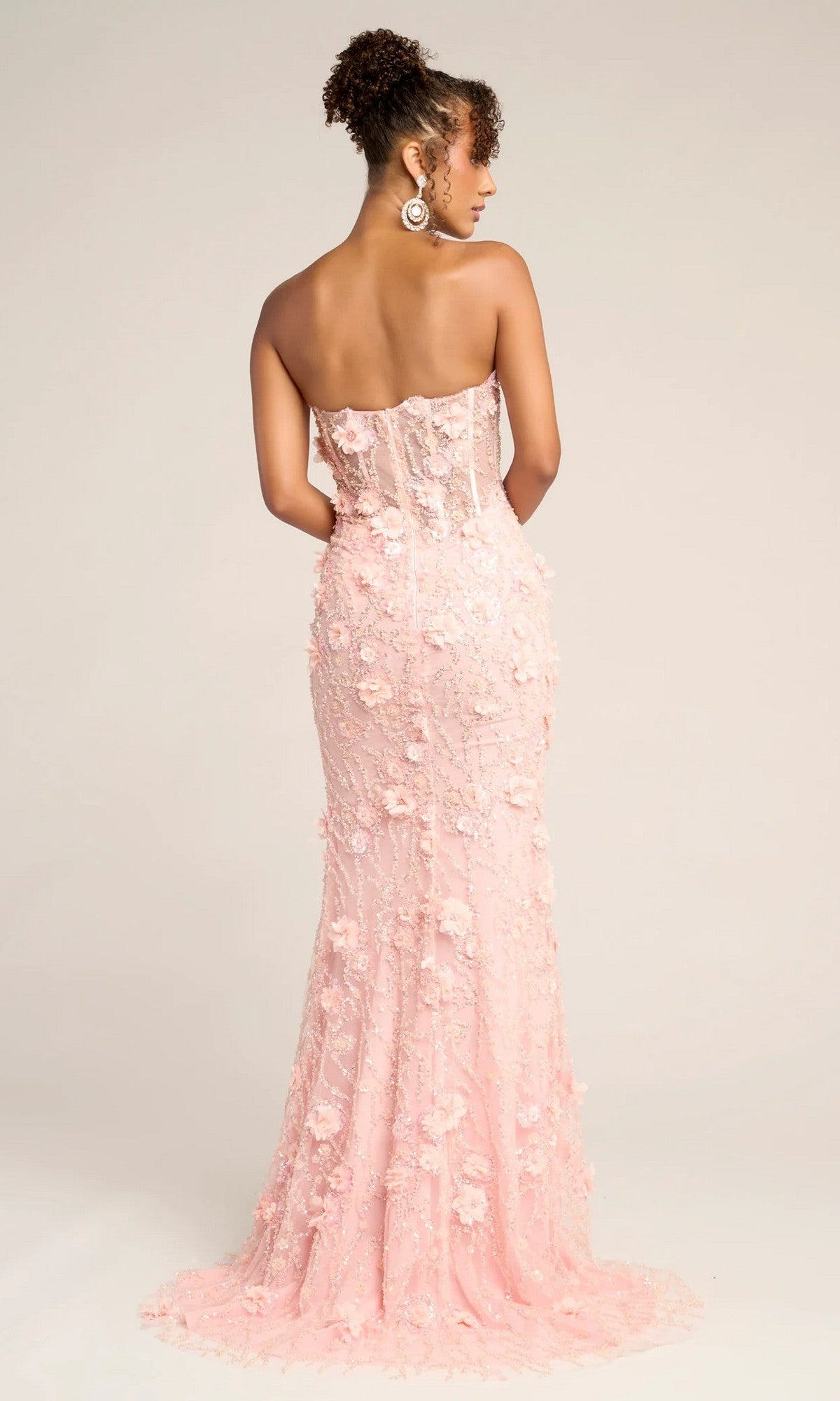 A woman faces away, wearing the Portia and Scarlett PS26011 long formal dress in light pink, featuring 3D flowers and delicate floral appliqué on a strapless, floor-length gown that radiates elegance.